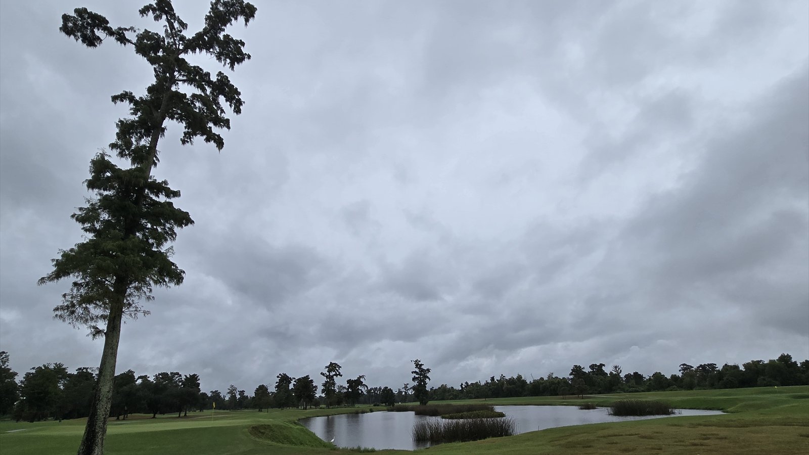 A green fairway curves beside a broad pond at TPC Louisiana beneath a partly cloudy sky.