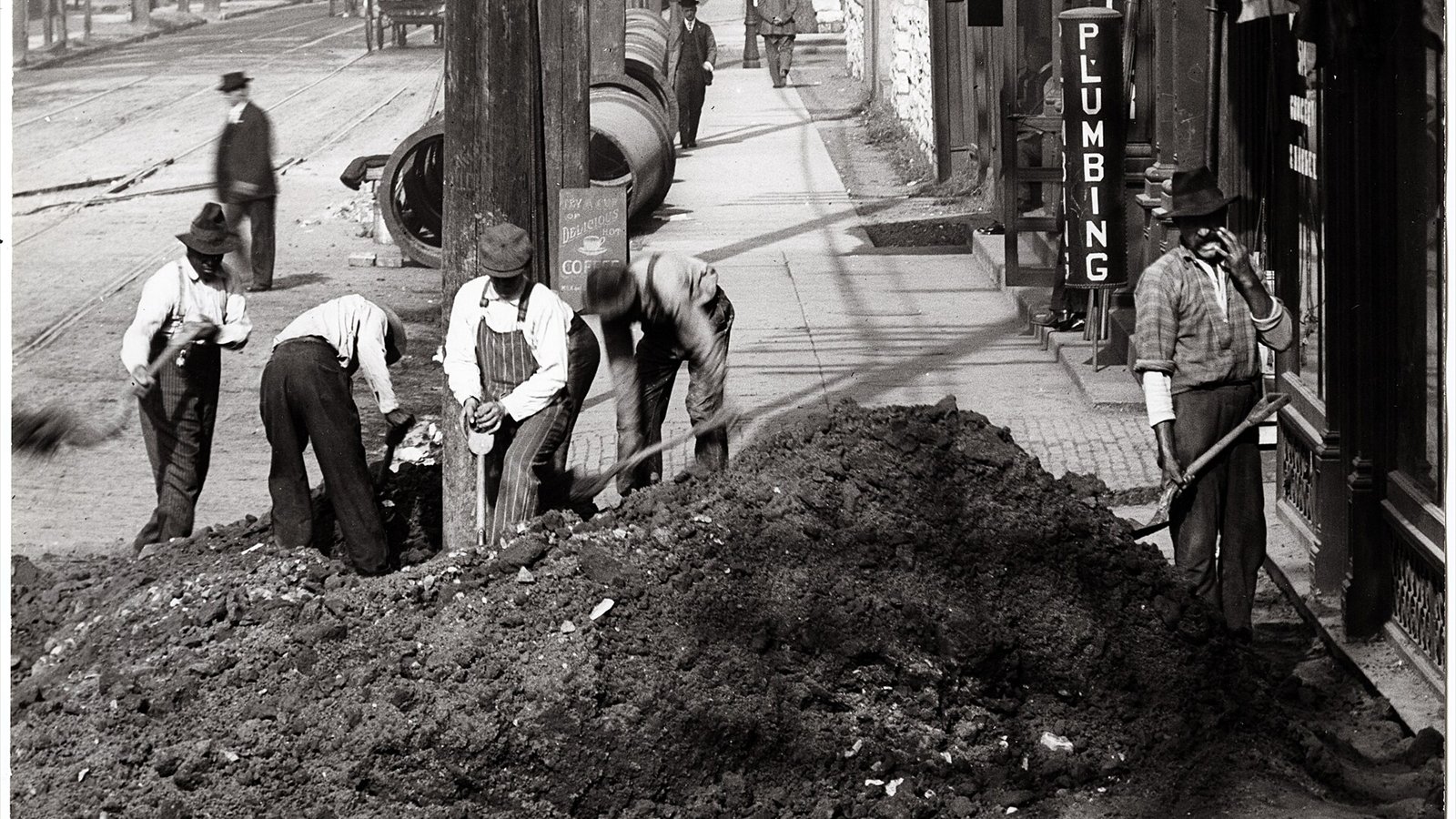 Horizontal, black and white photograph showing a street crew backfilling after laying new underground utilities on Sarah Avenue near Washington Boulevard. A mound of dirt sits in the foreground, covering the sidewalk…