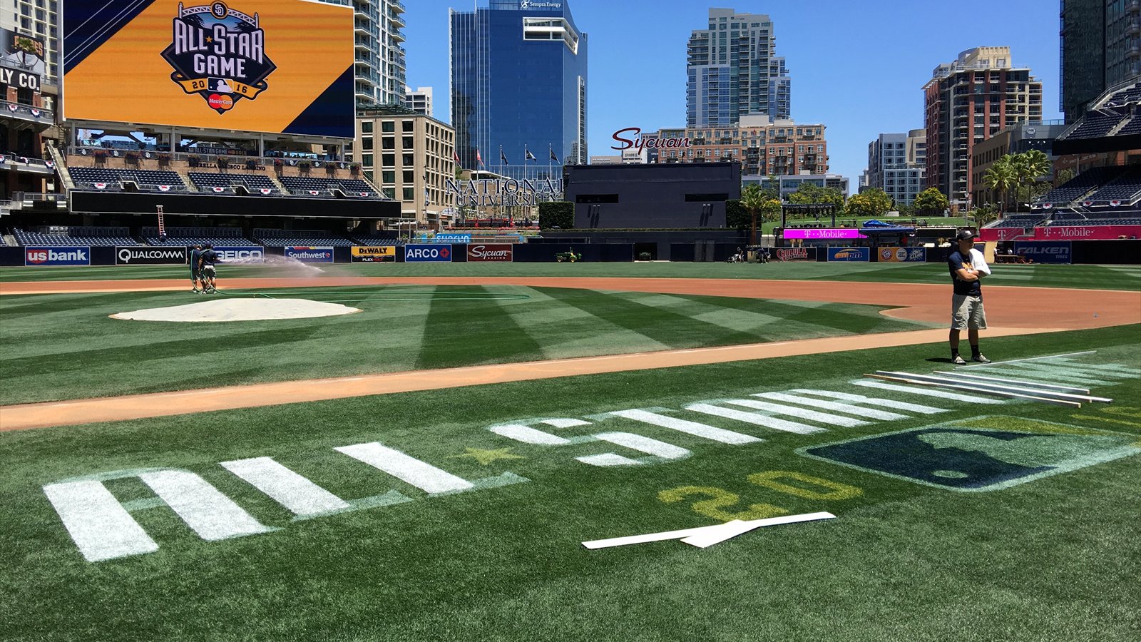 Petco Park's outfield and downtown San Diego skyline under a clear blue sky while a grounds crew waters the field.