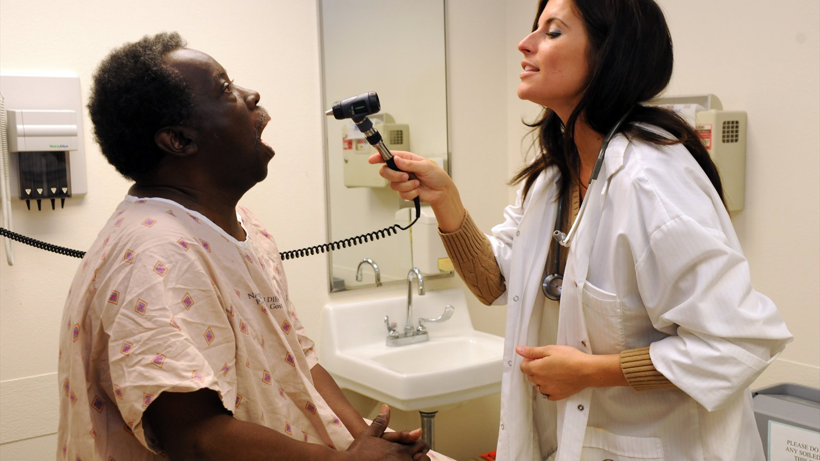 A nurse practitioner uses a handheld scope to examine a seated patient's open mouth in a clinic room.