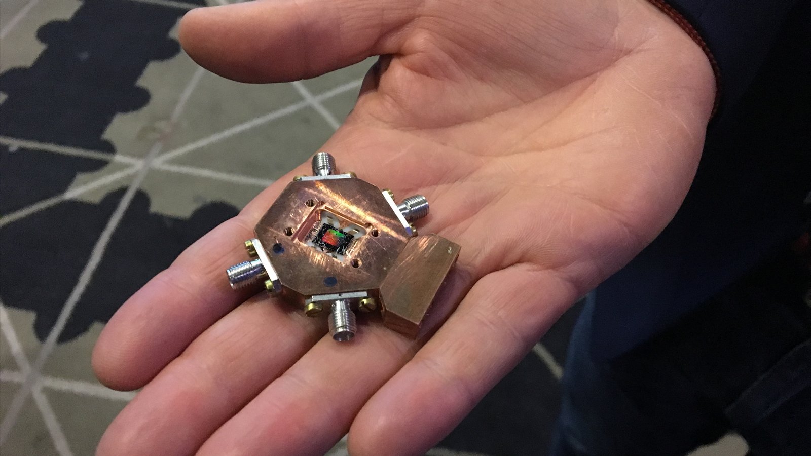 A hand holds a small copper-colored quantum processor with metal connectors arranged around its edges.
