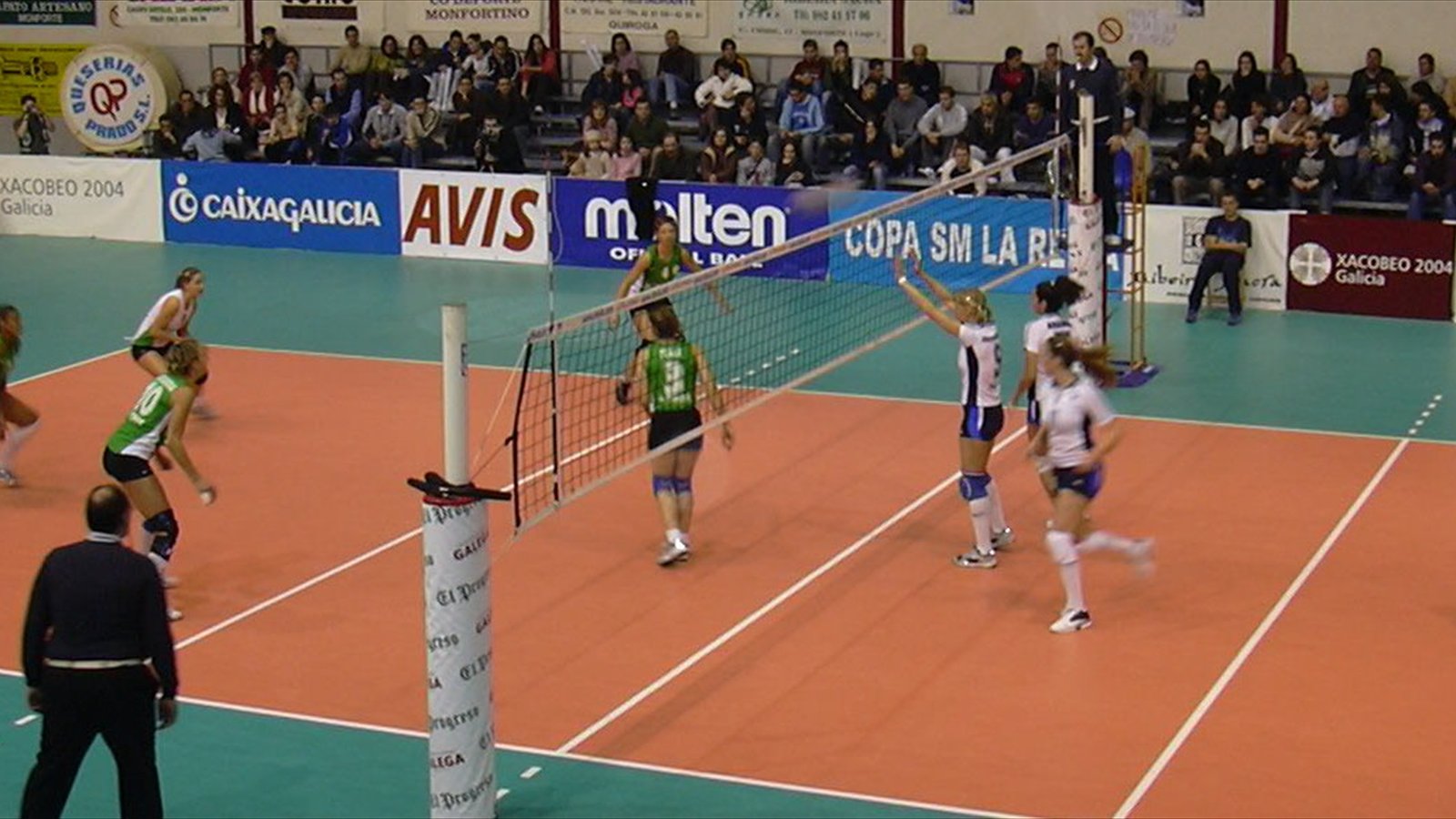 Two women's volleyball teams contest the ball at the net on an indoor court as spectators watch from the stands.