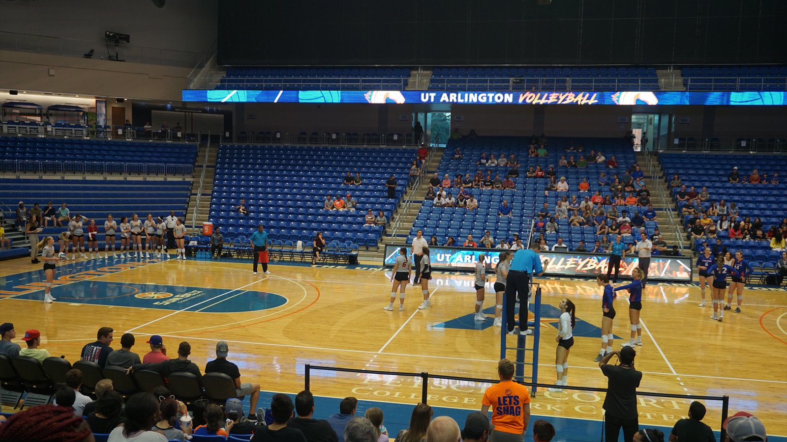 Two college volleyball teams position themselves on opposite sides of the net during a match in a crowded gym.