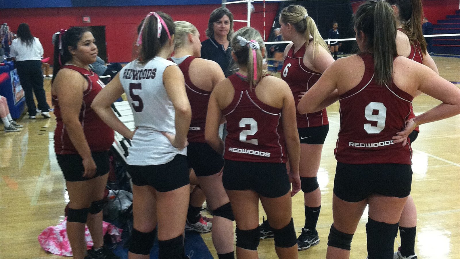 College of the Redwoods women's volleyball players gather around a coach beside an indoor court.