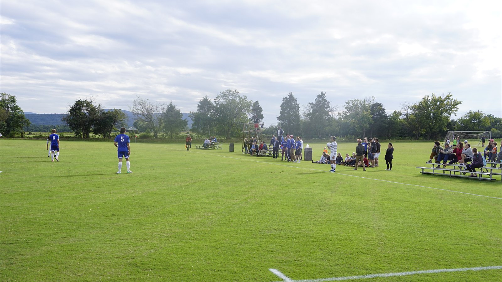College men's soccer players stand across a grass field while teammates and spectators watch from the sideline.
