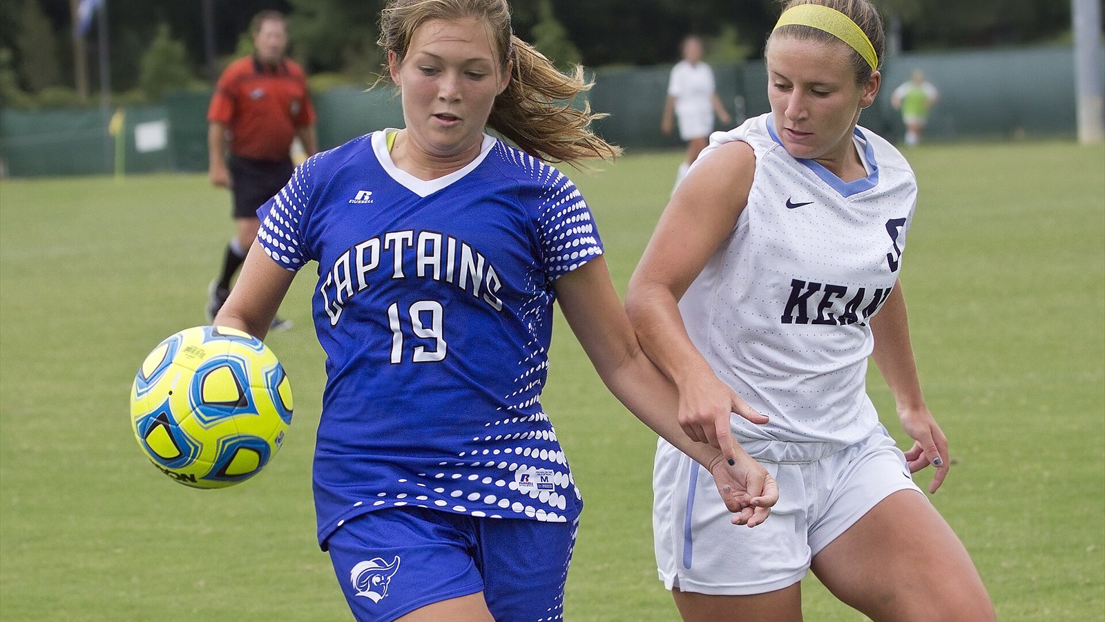 Women's college soccer players run toward the ball during a match on a green field.