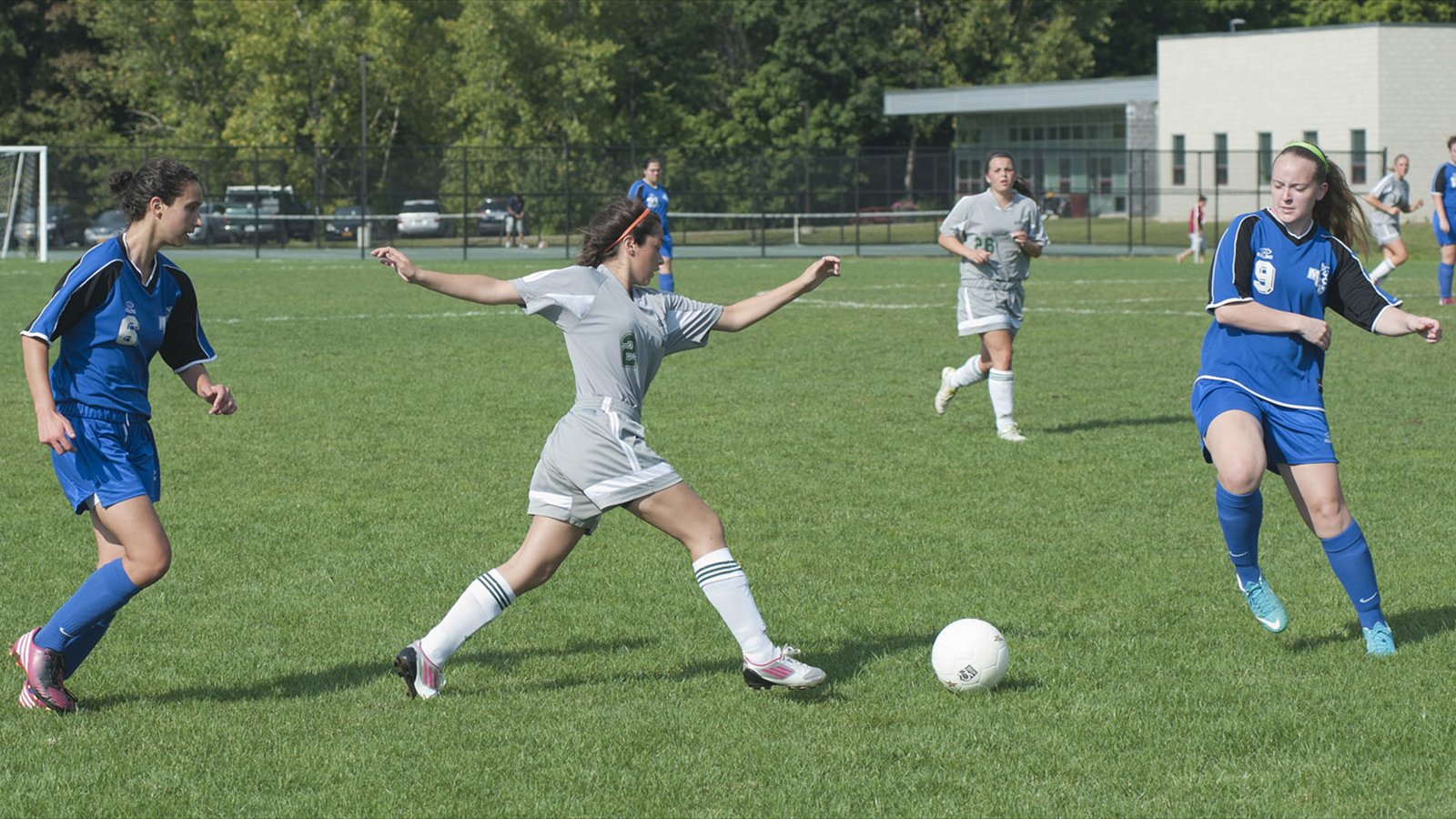 Women's college soccer players converge on the ball on an outdoor field.