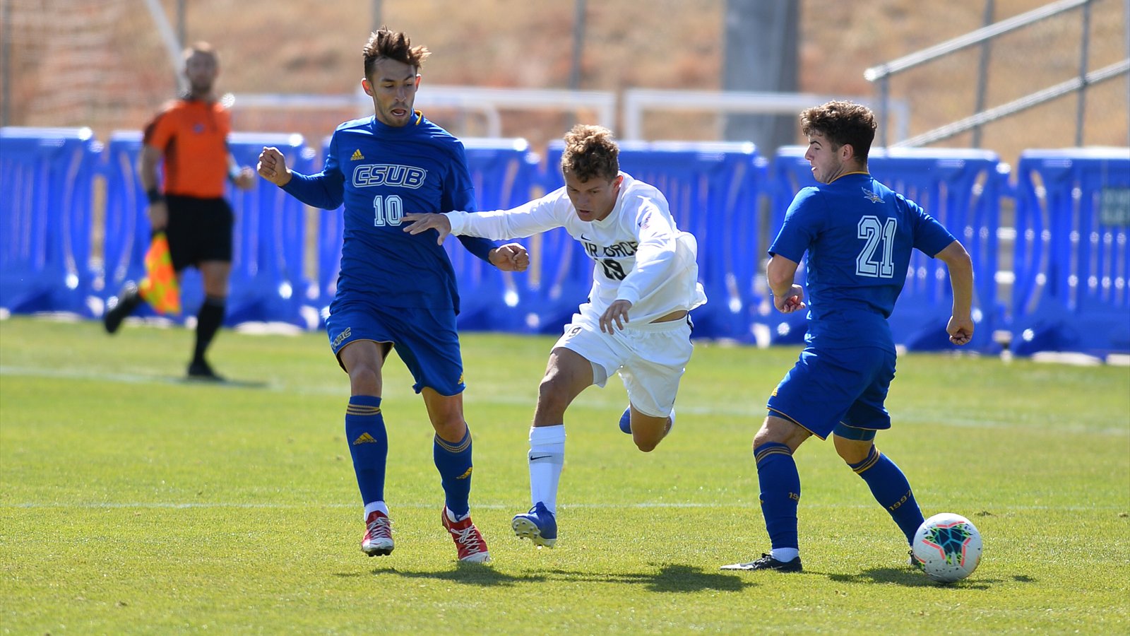 Two Cal State Bakersfield players in blue close around an Air Force player in white who is chasing the ball on a sunny soccer field.