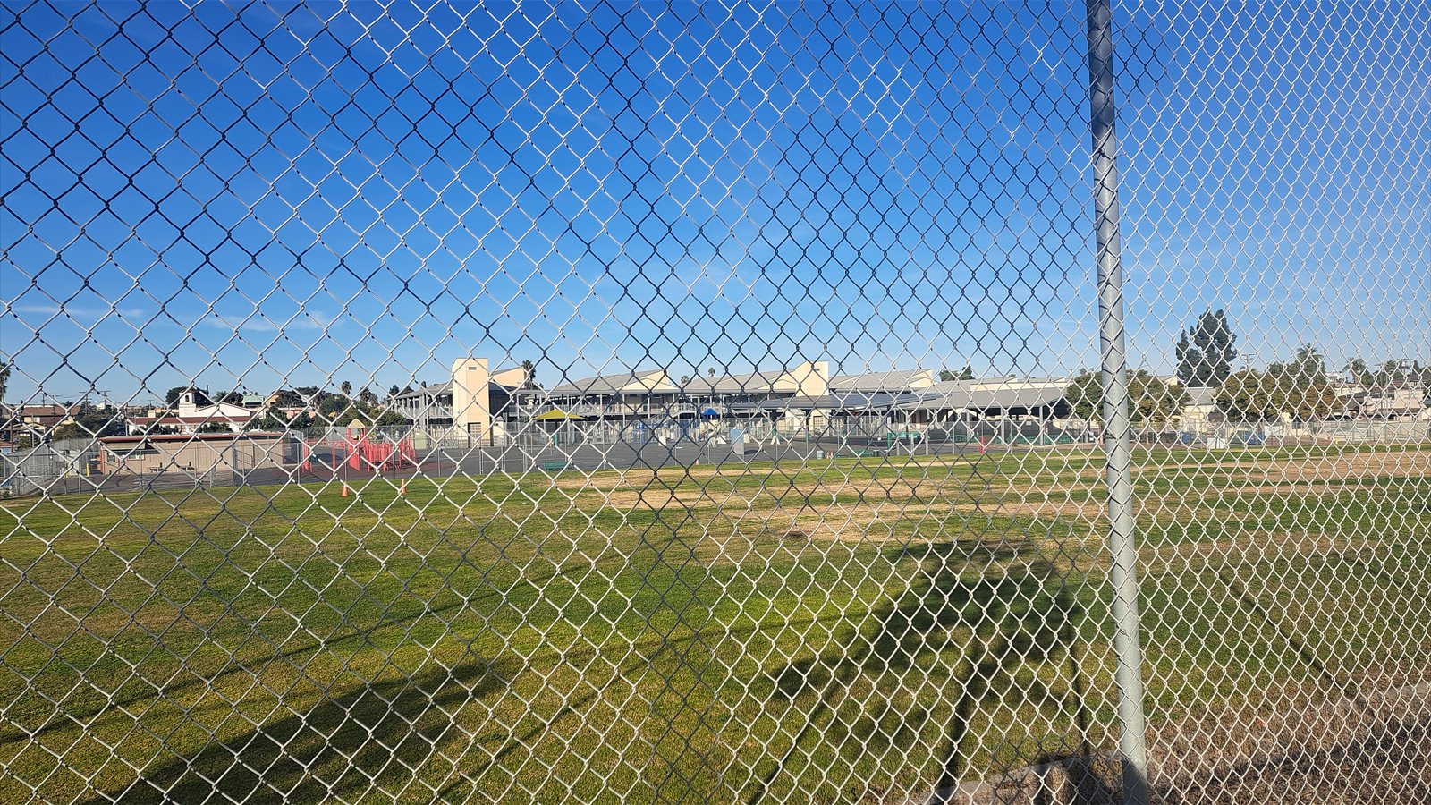A chain-link fence frames a grassy athletic field and elementary school buildings under a clear blue sky.