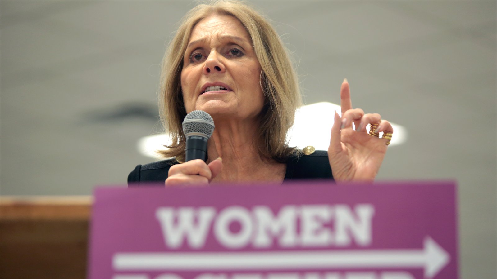 Gloria Steinem speaks into a handheld microphone and raises one finger behind a Women Together sign.