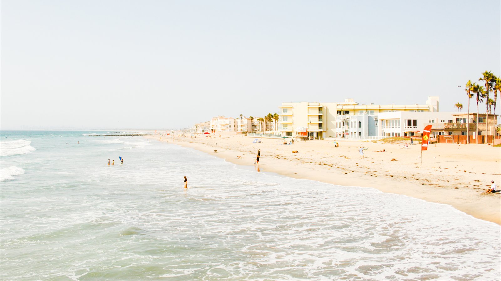 A bright view from the Imperial Beach Pier across surf, sand, beachgoers and low-rise waterfront buildings.