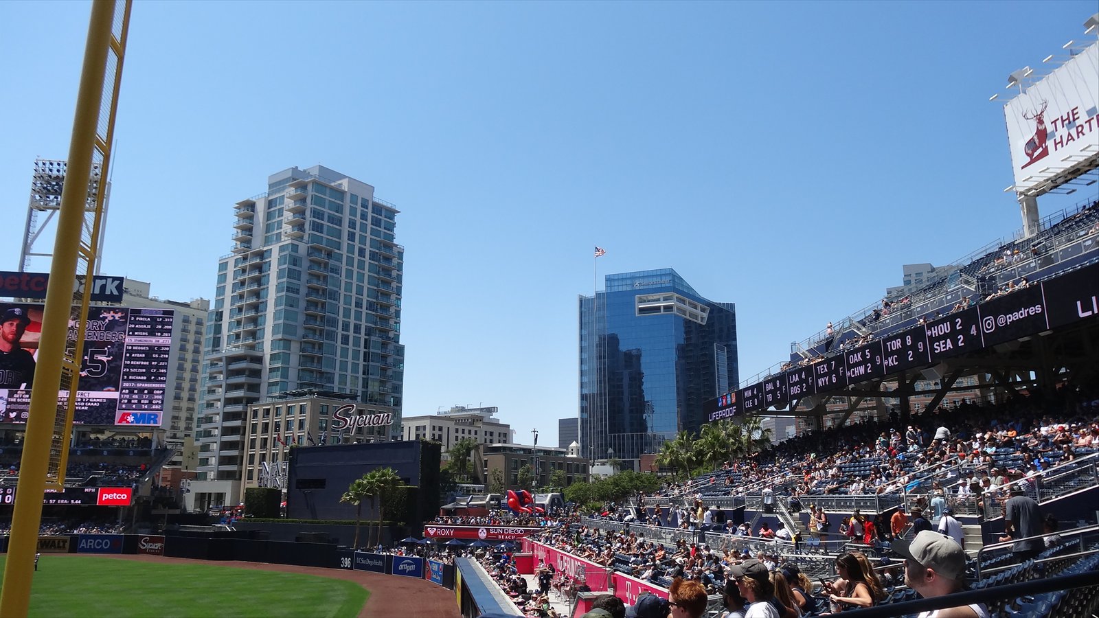 A yellow foul pole rises beside Petco Park's outfield, with occupied seats, scoreboards and downtown buildings under a clear sky.