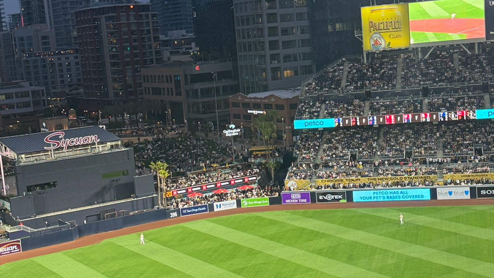 A baseball game at Petco Park is viewed across the outfield toward an illuminated scoreboard and downtown towers.