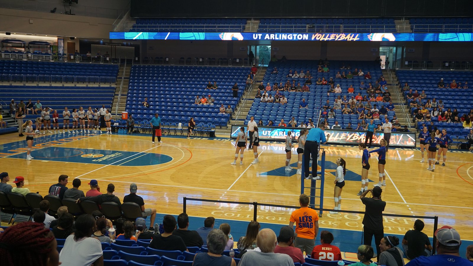 Two college volleyball teams position themselves on opposite sides of the net during a match in a crowded gym.