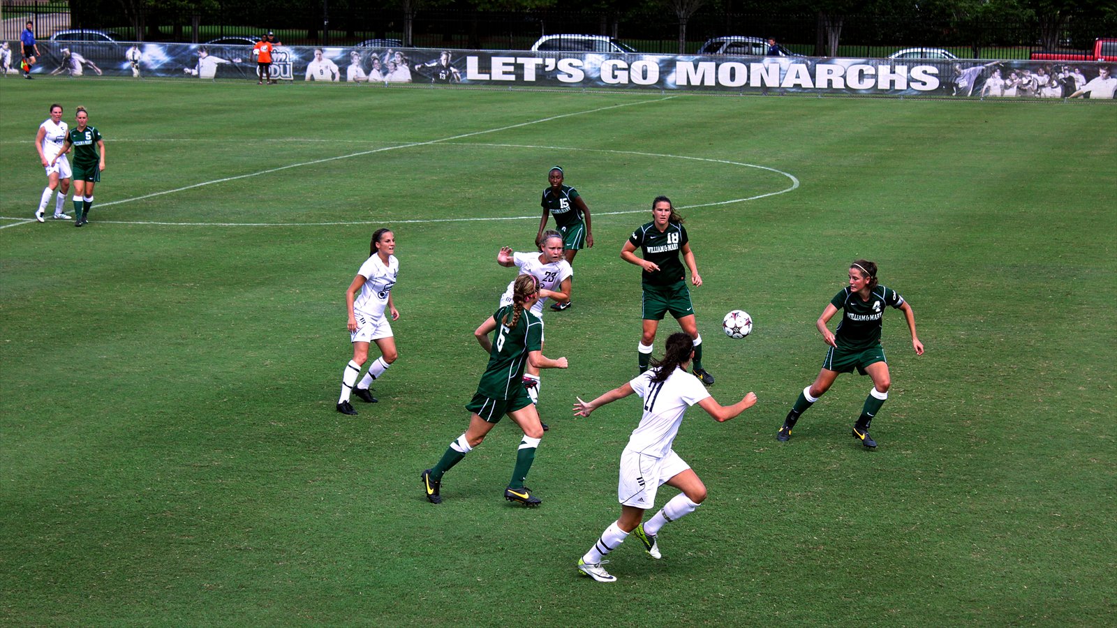 Women's college soccer players run toward the ball on a grass field during a match.