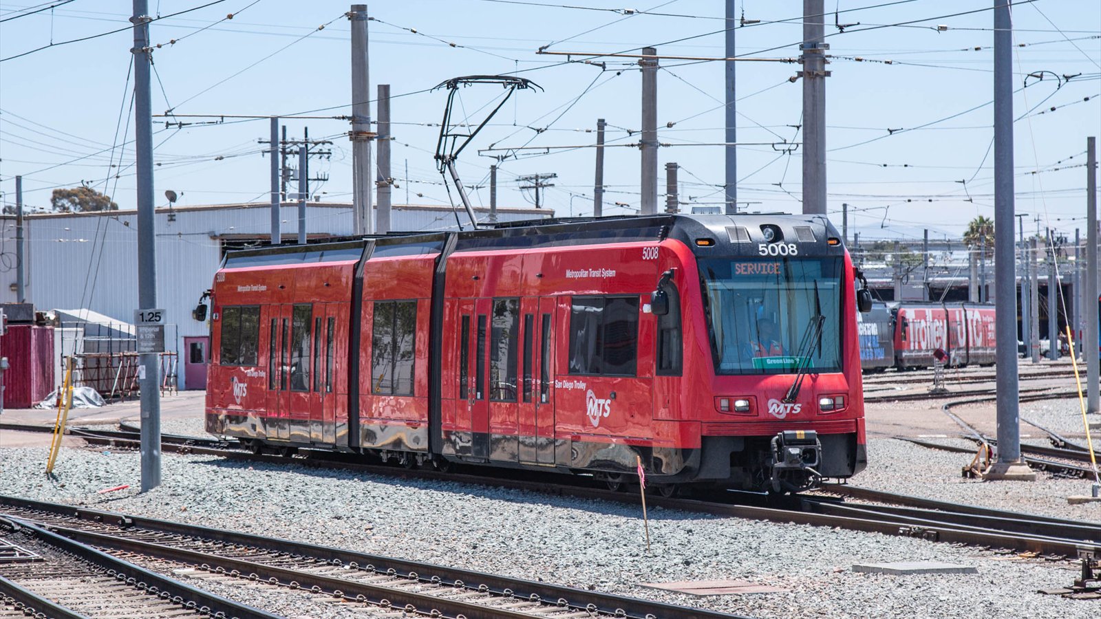 A red San Diego MTS trolley moves through a rail yard beneath overhead electrical wires.