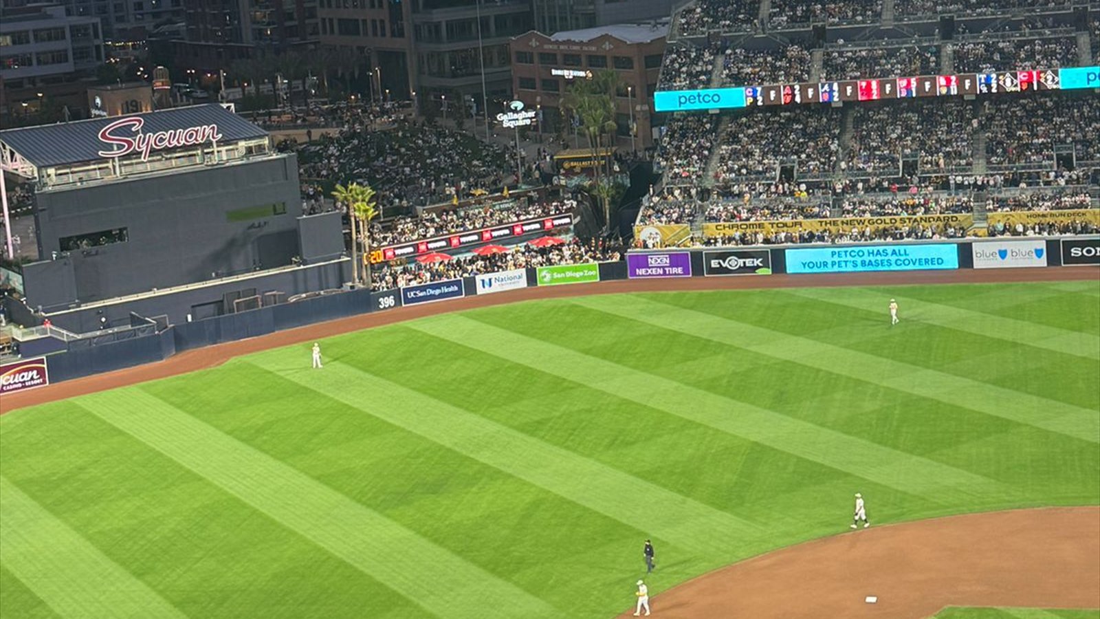 A baseball game at Petco Park viewed across the outfield toward the lit scoreboard and downtown high-rises.