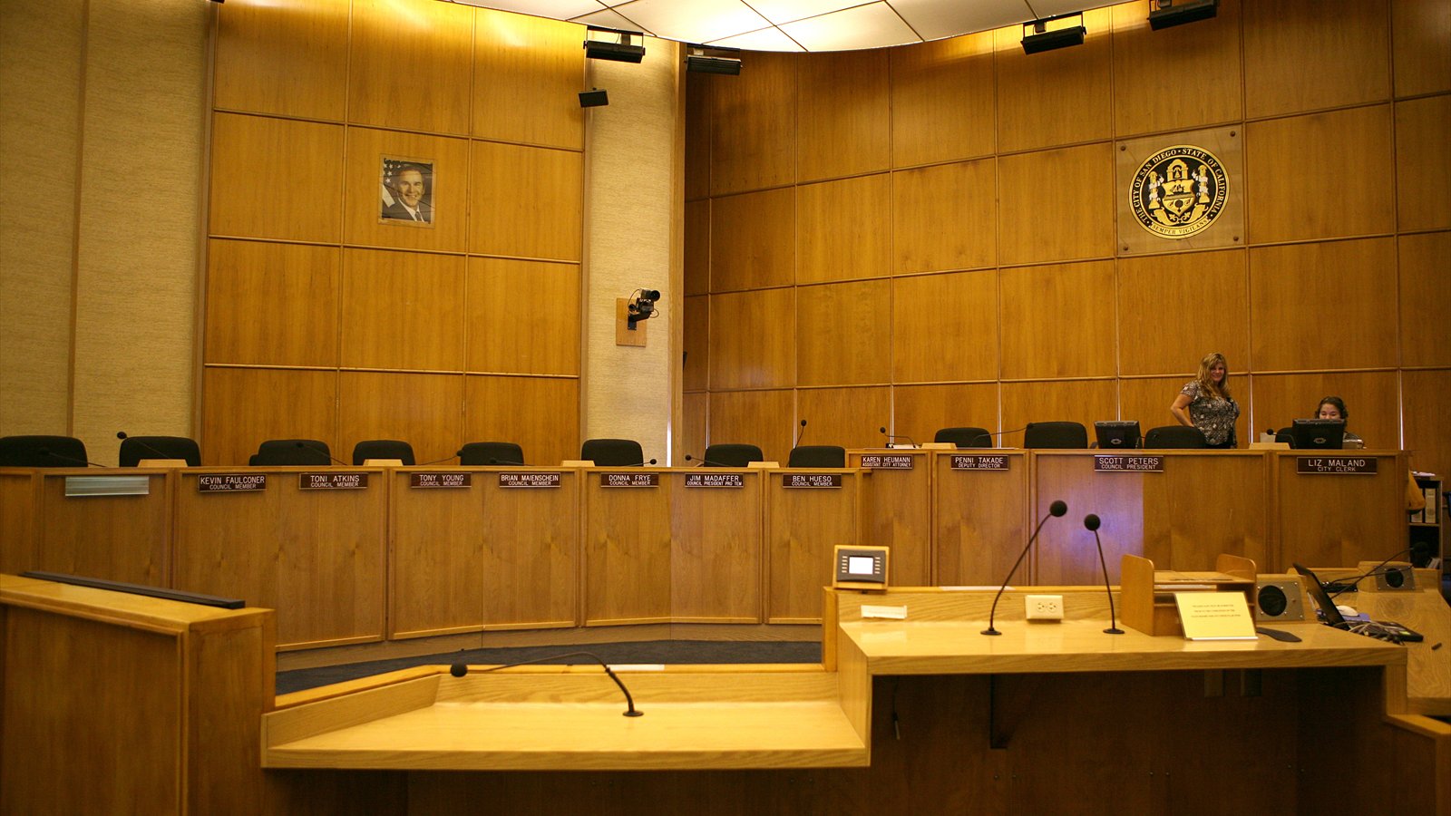 The wood-paneled San Diego City Council chambers contain a curved dais, empty chairs and microphones.