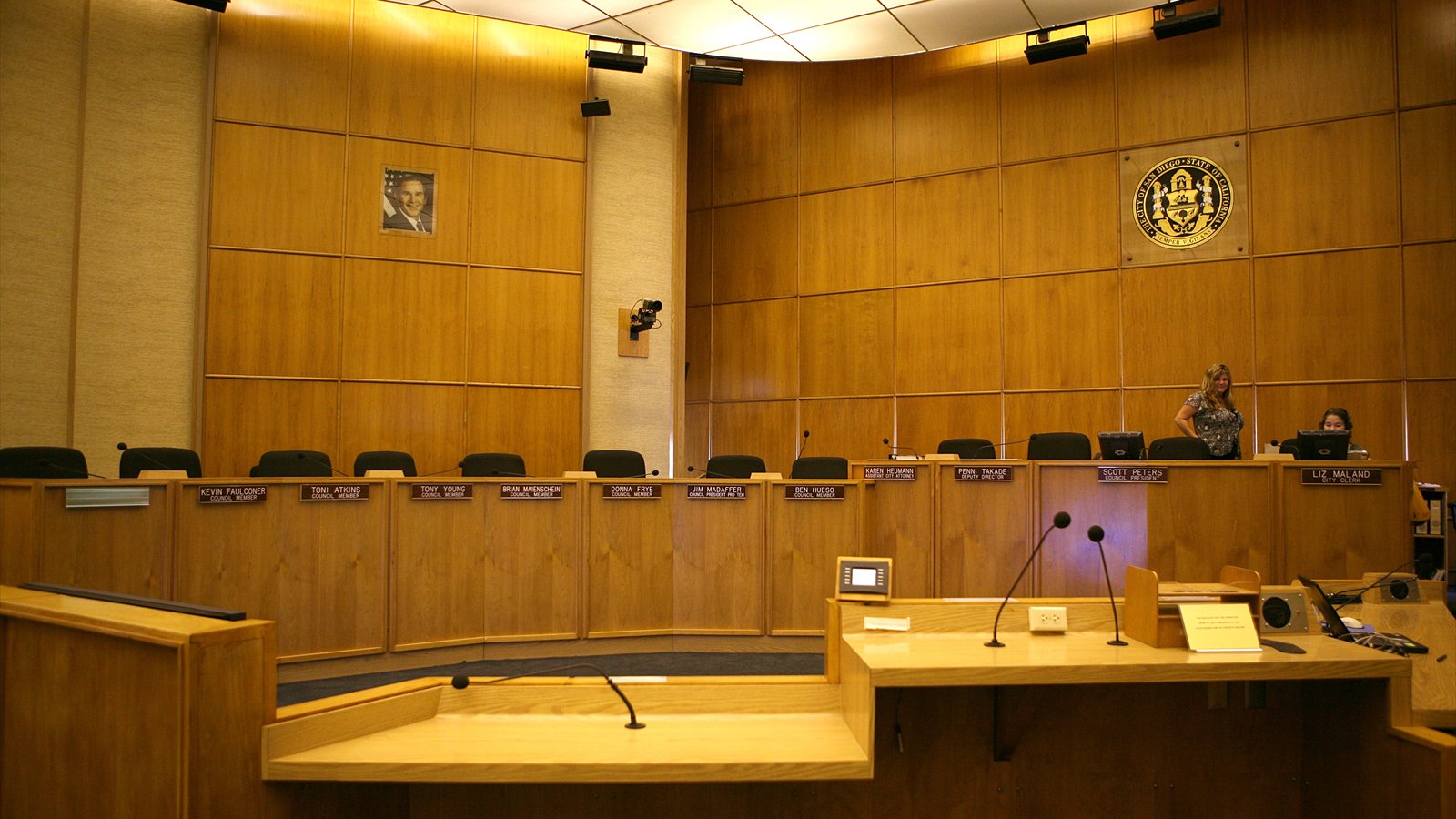 Curved wood desks and microphones face the City of San Diego seal inside the council chamber.