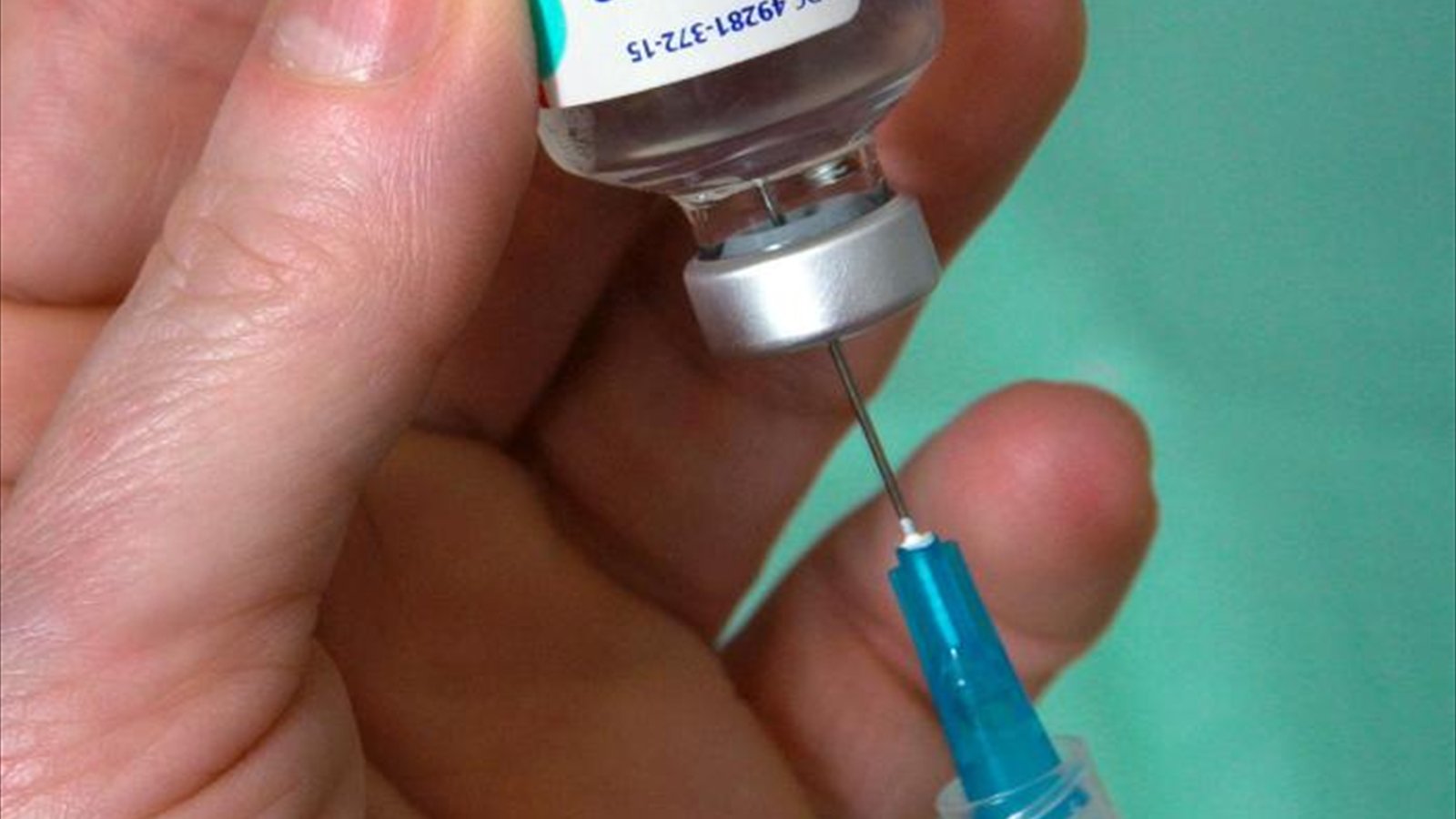 Hands hold a small influenza-vaccine vial as a syringe needle enters its stopper.