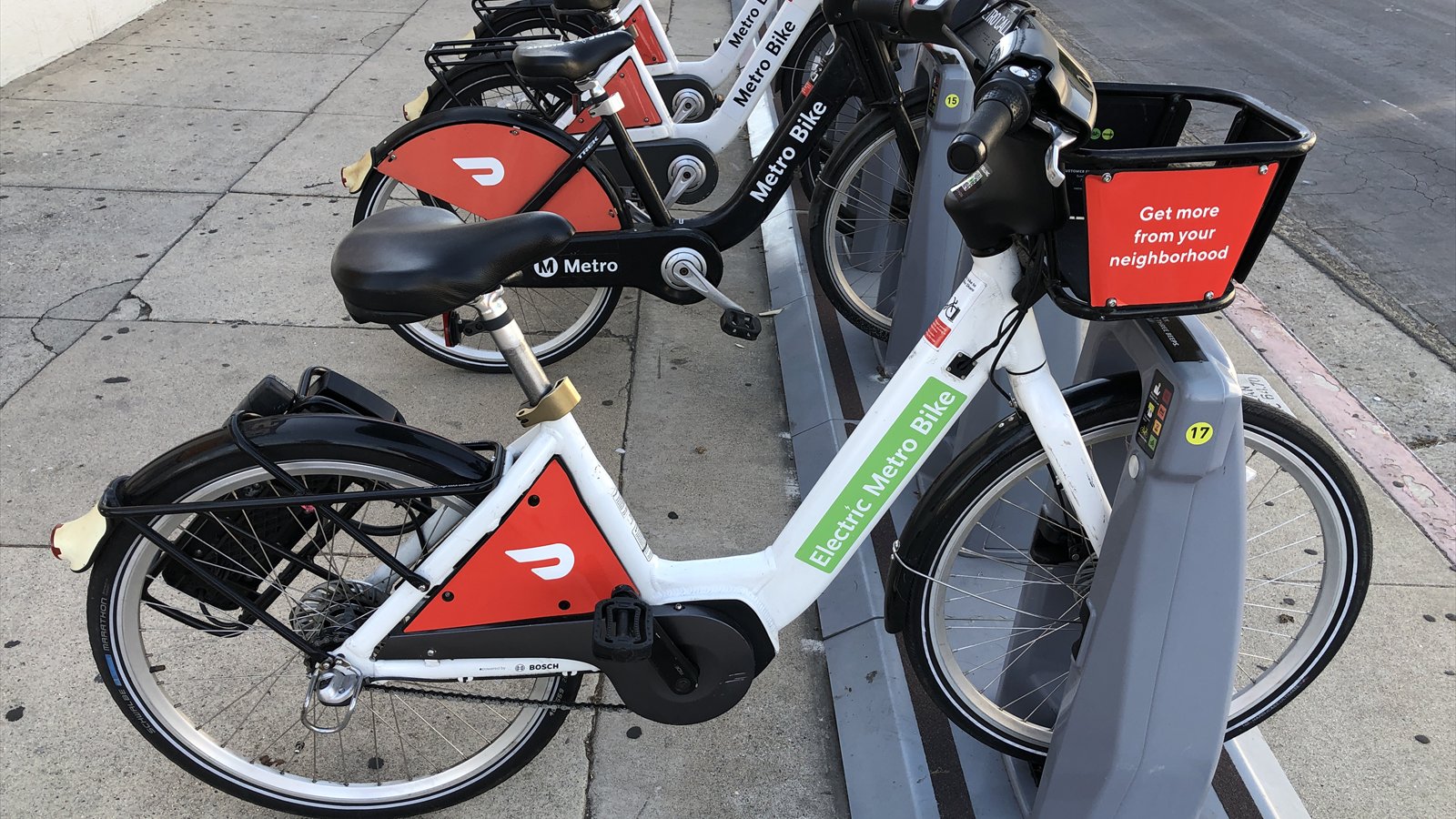 A white electric bike-share bicycle is docked with several red and black rental bikes on a paved sidewalk.