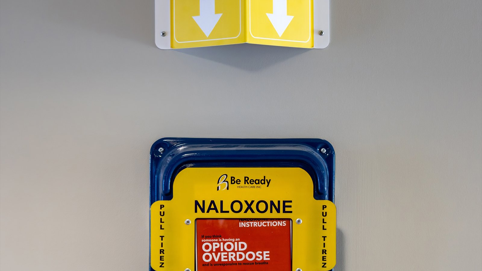 A yellow emergency naloxone box mounted on a wall beneath an opioid-overdose sign.