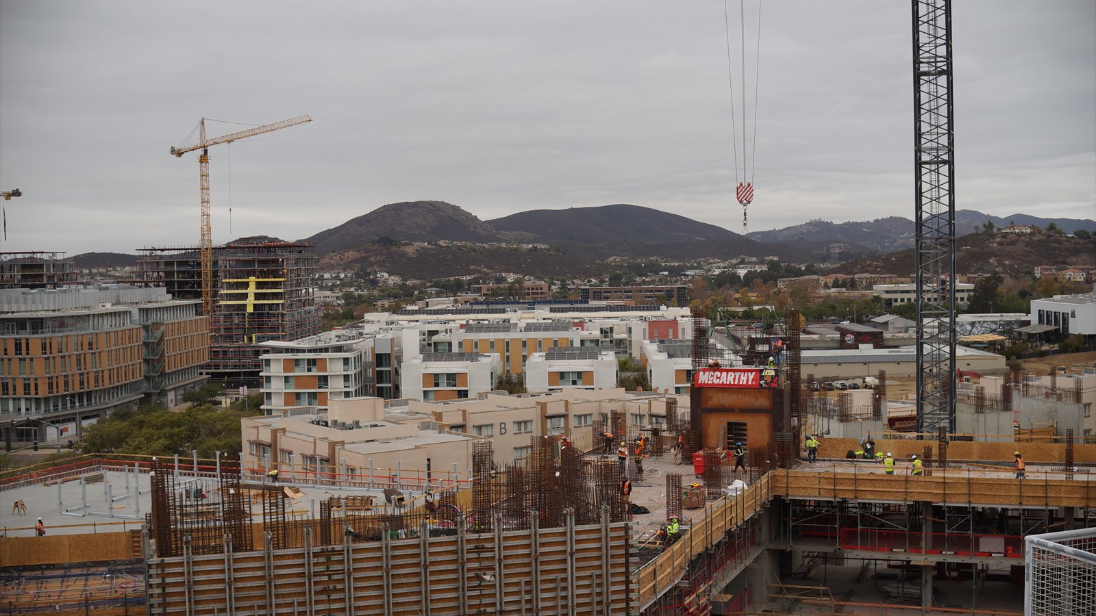Cranes and workers surround multistory housing under construction near Cal State San Marcos, with hills in the distance.