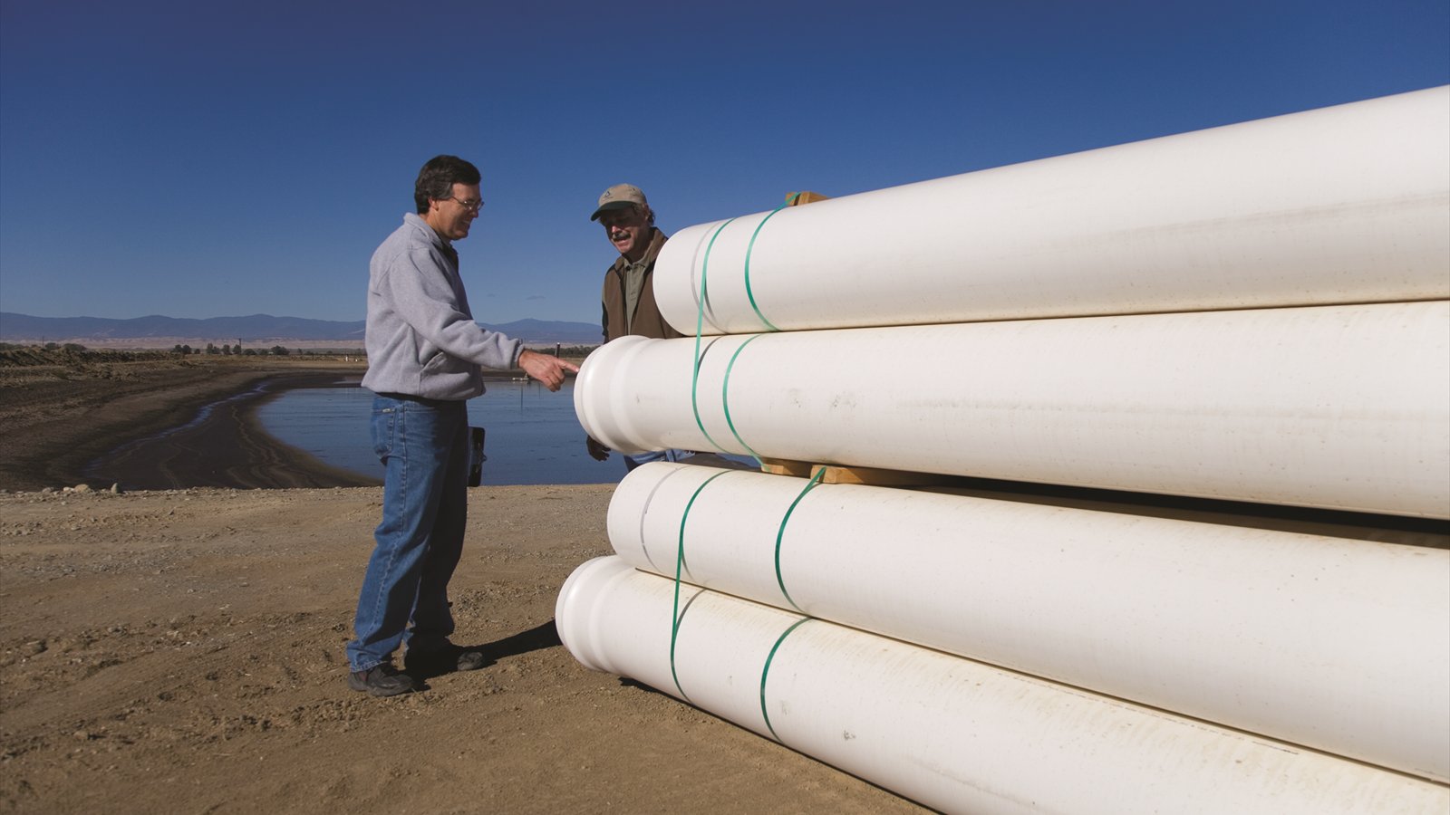 Engineers stand beside large sections of black plastic pipe laid across a California field.