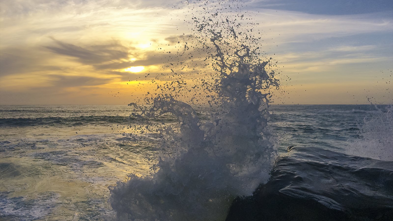 A wave bursts upward against dark rocks at Windansea Beach as the sun sets over the Pacific.