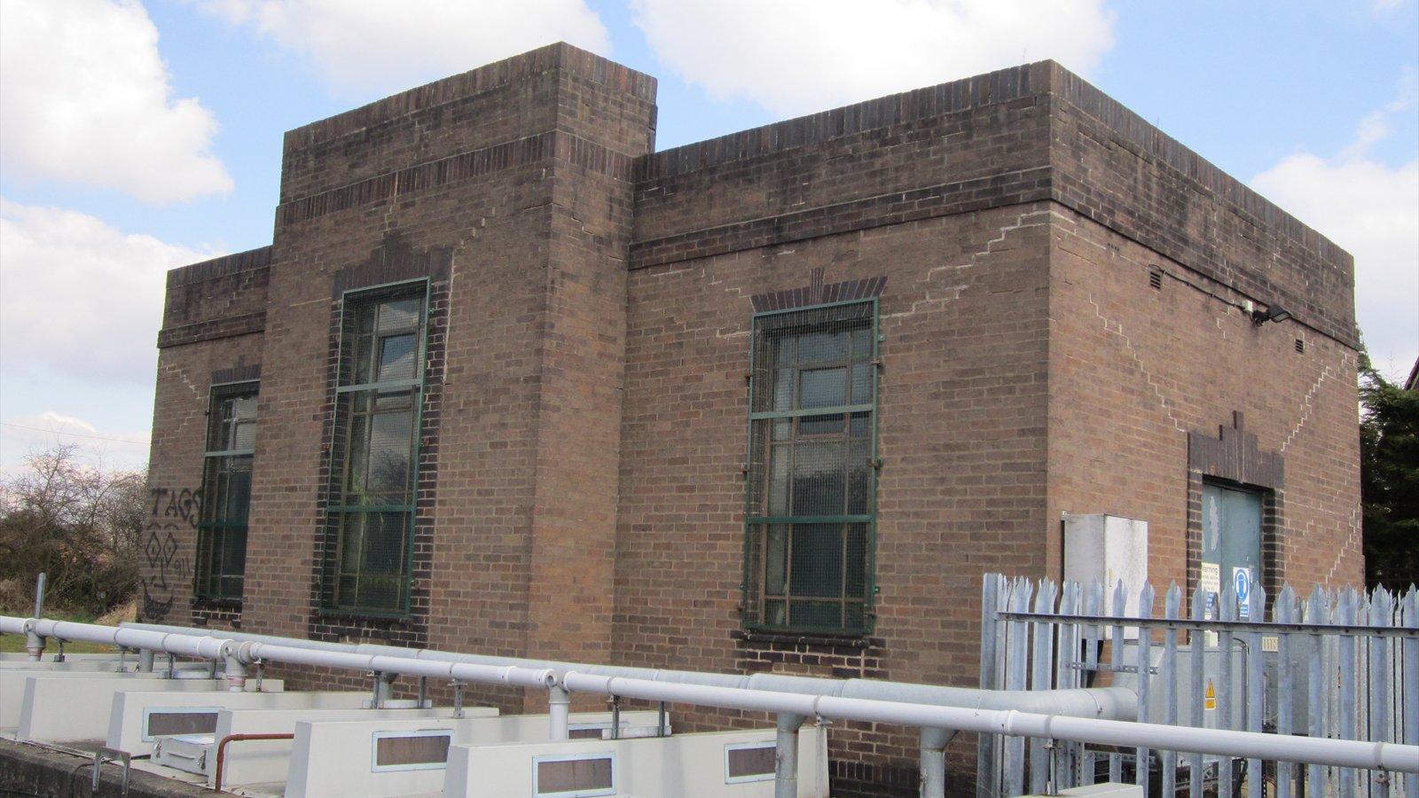 A brick wastewater pumping station stands behind a metal fence beside a paved road.