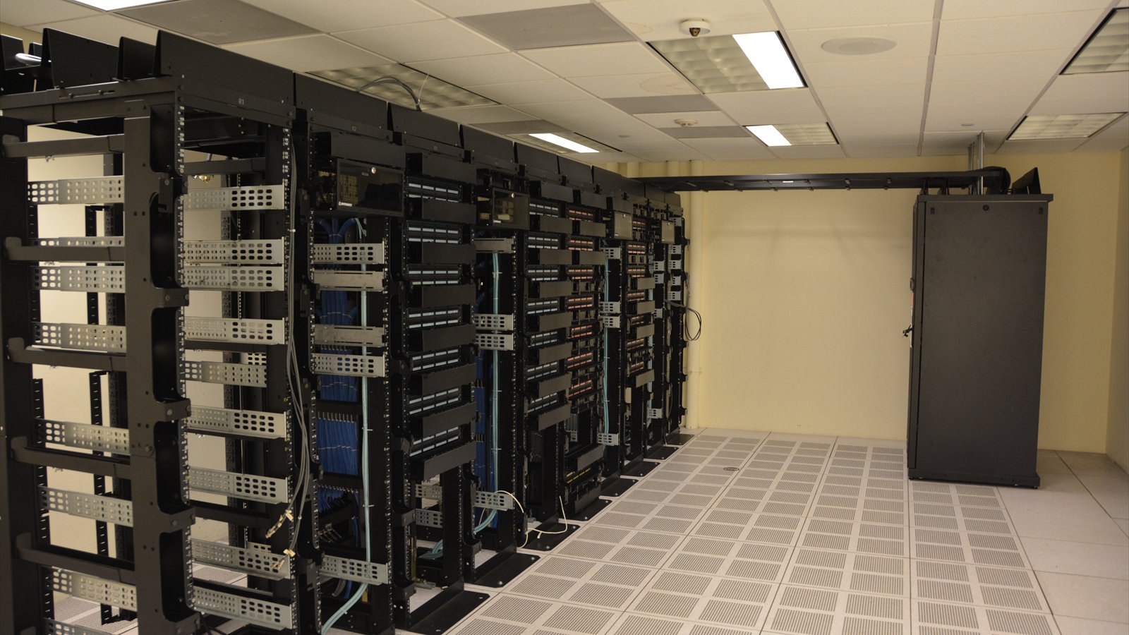 Rows of black server racks stand beneath overhead cables in a data center.