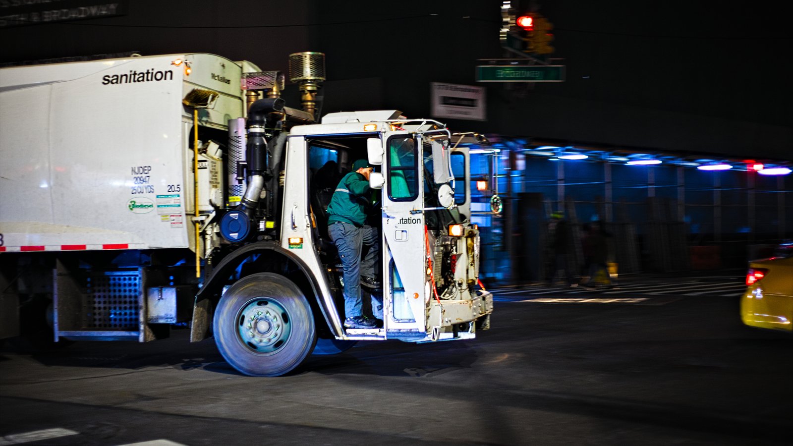 A white sanitation collection truck travels along a city street beside parked cars.