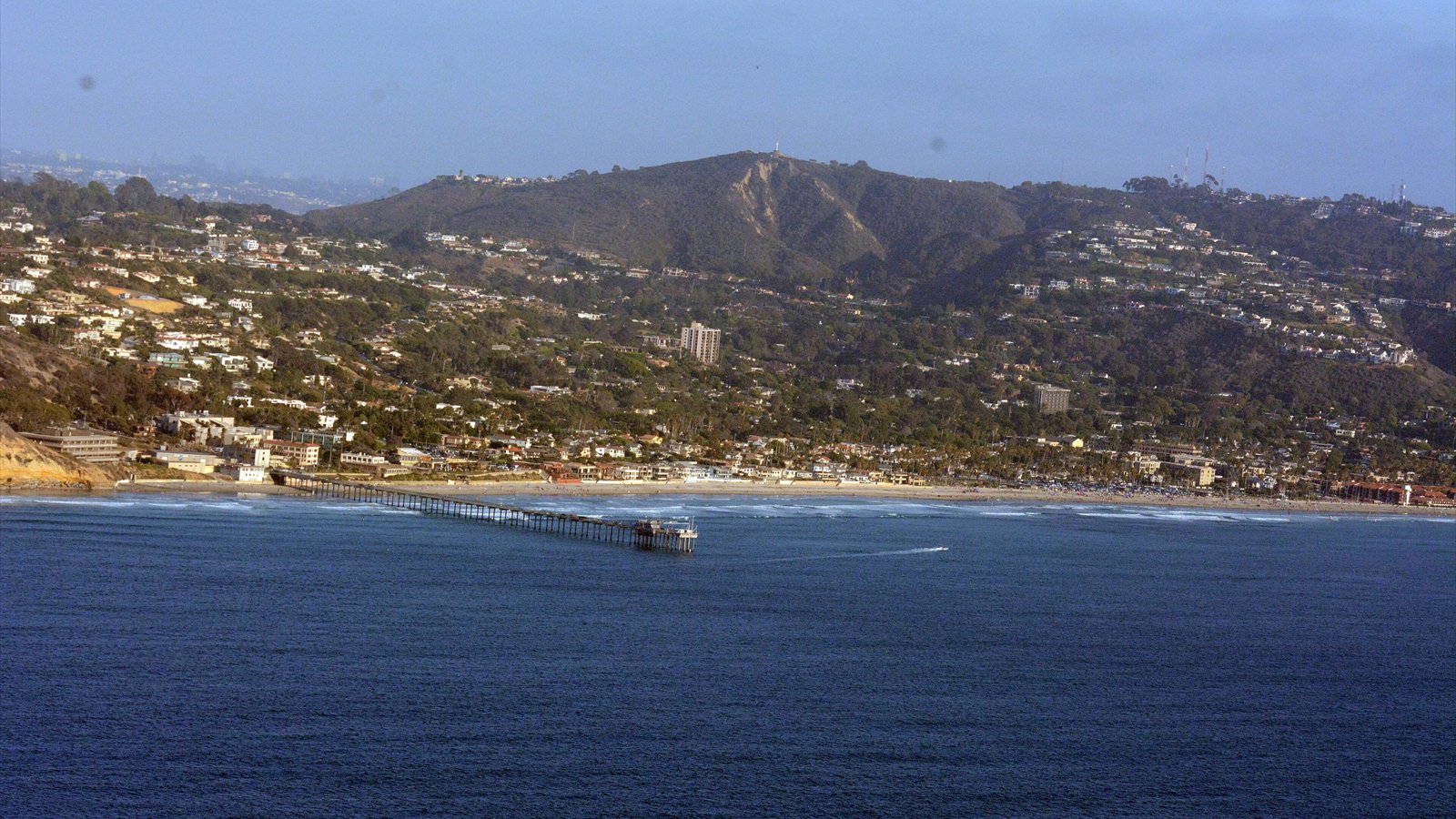The Scripps Pier extends from the La Jolla shoreline over blue ocean water beneath a clear sky.
