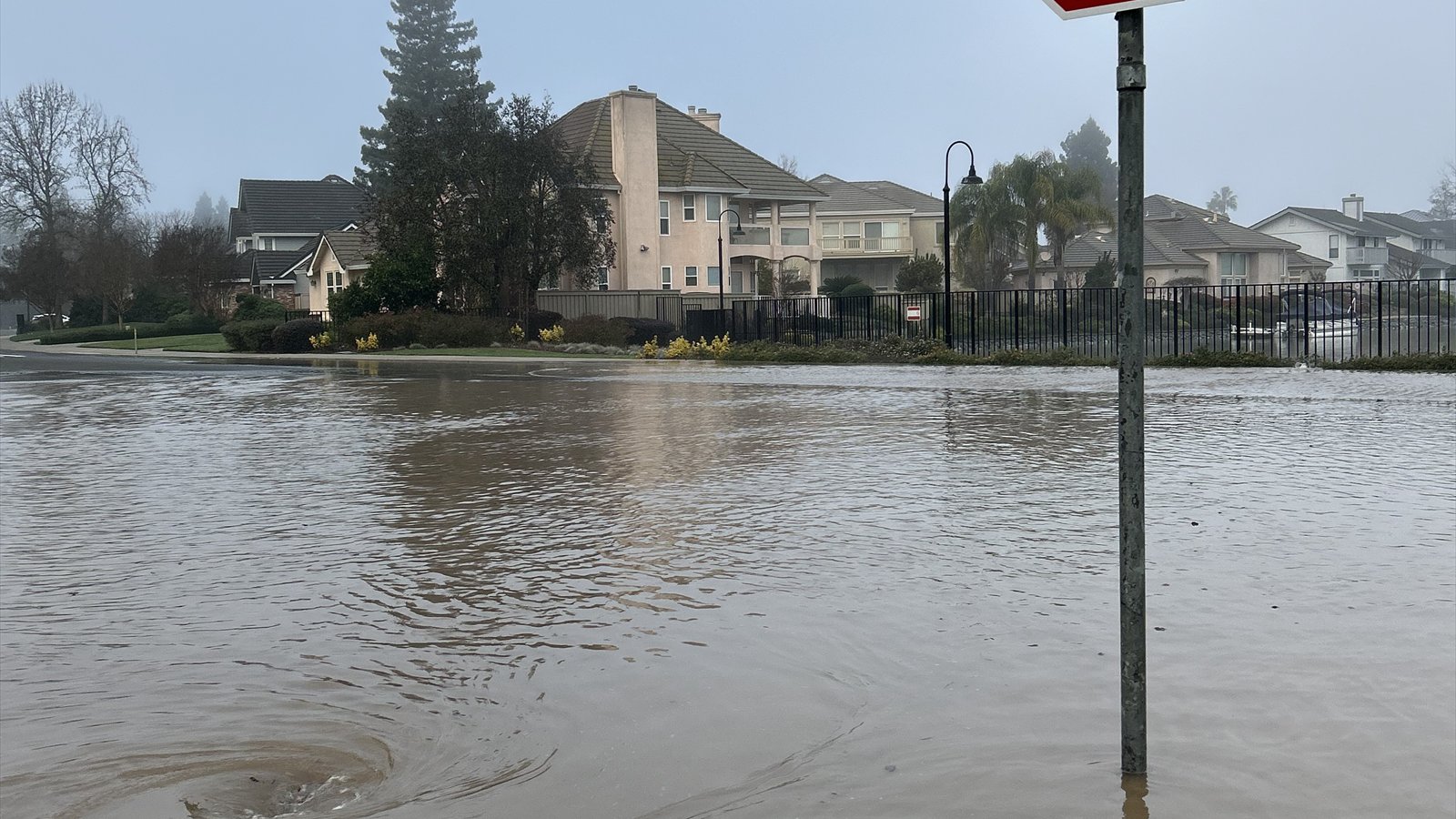 Brown floodwater covers a Sacramento residential street, with a sign pole and a whirlpool in the foreground.