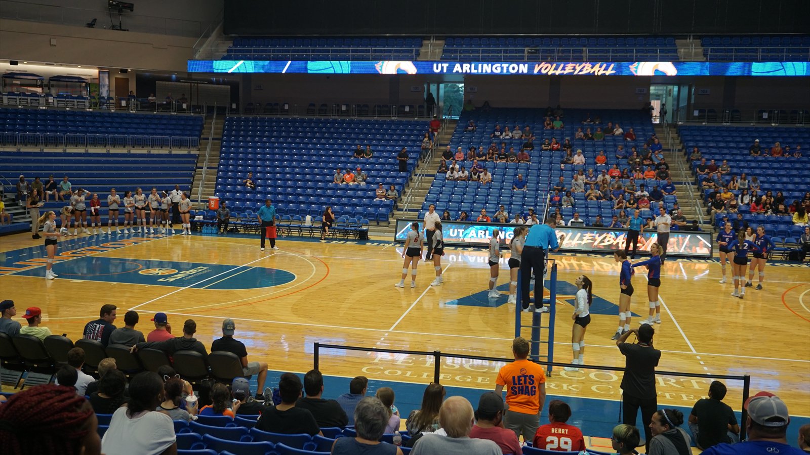 Two indoor volleyball teams defend at the net during a college match as the ball crosses above them.