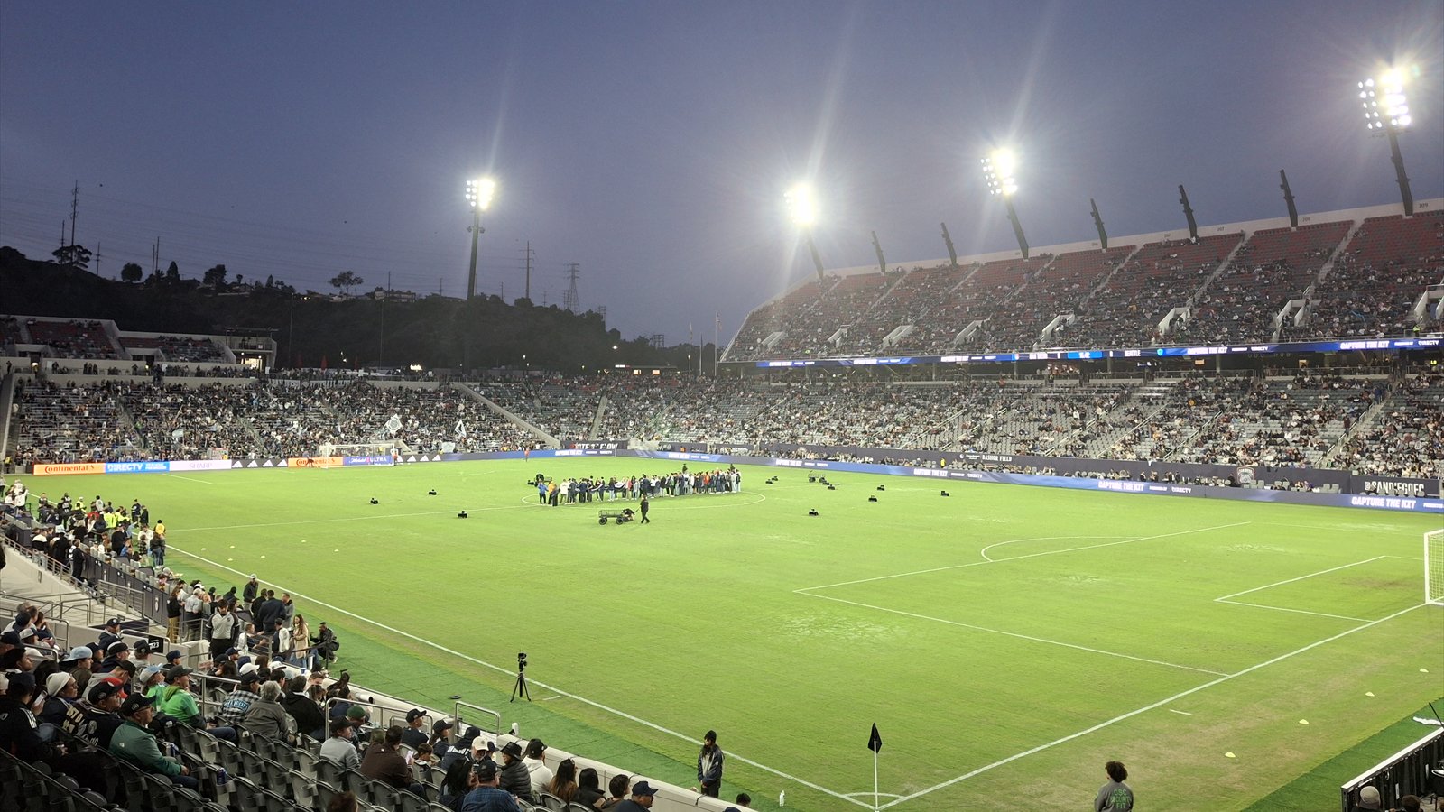 Spectators sit beneath bright stadium lights around the green field at Snapdragon Stadium during an evening event.