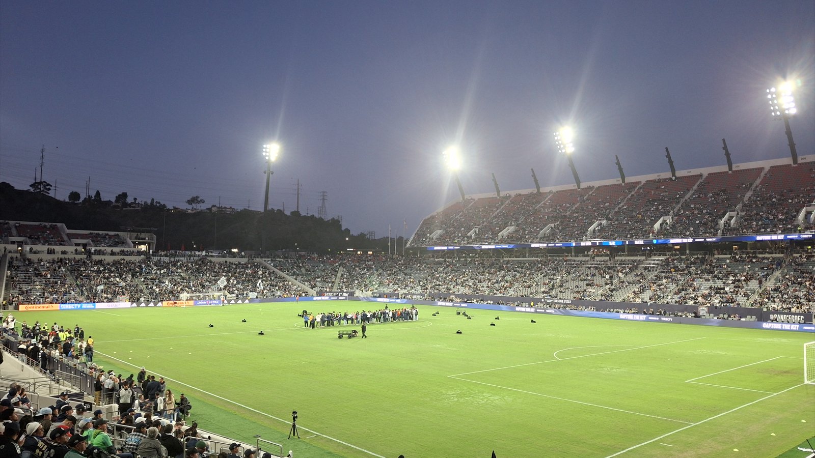 Spectators sit beneath bright stadium lights around the green field at Snapdragon Stadium during an evening event.