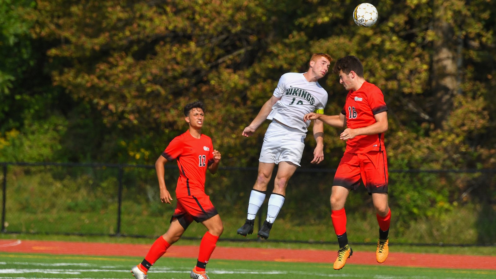 Three men's college soccer players watch the ball after one jumps for a header on a grass field.