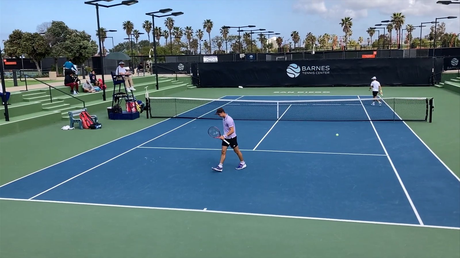 Two players change ends on a blue-and-green hard court at the Barnes Tennis Center as an official watches from the chair.