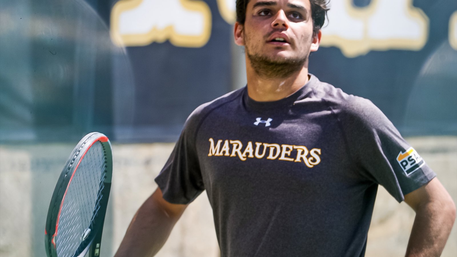 A men's college tennis player holds a racquet while standing on an outdoor hard court.