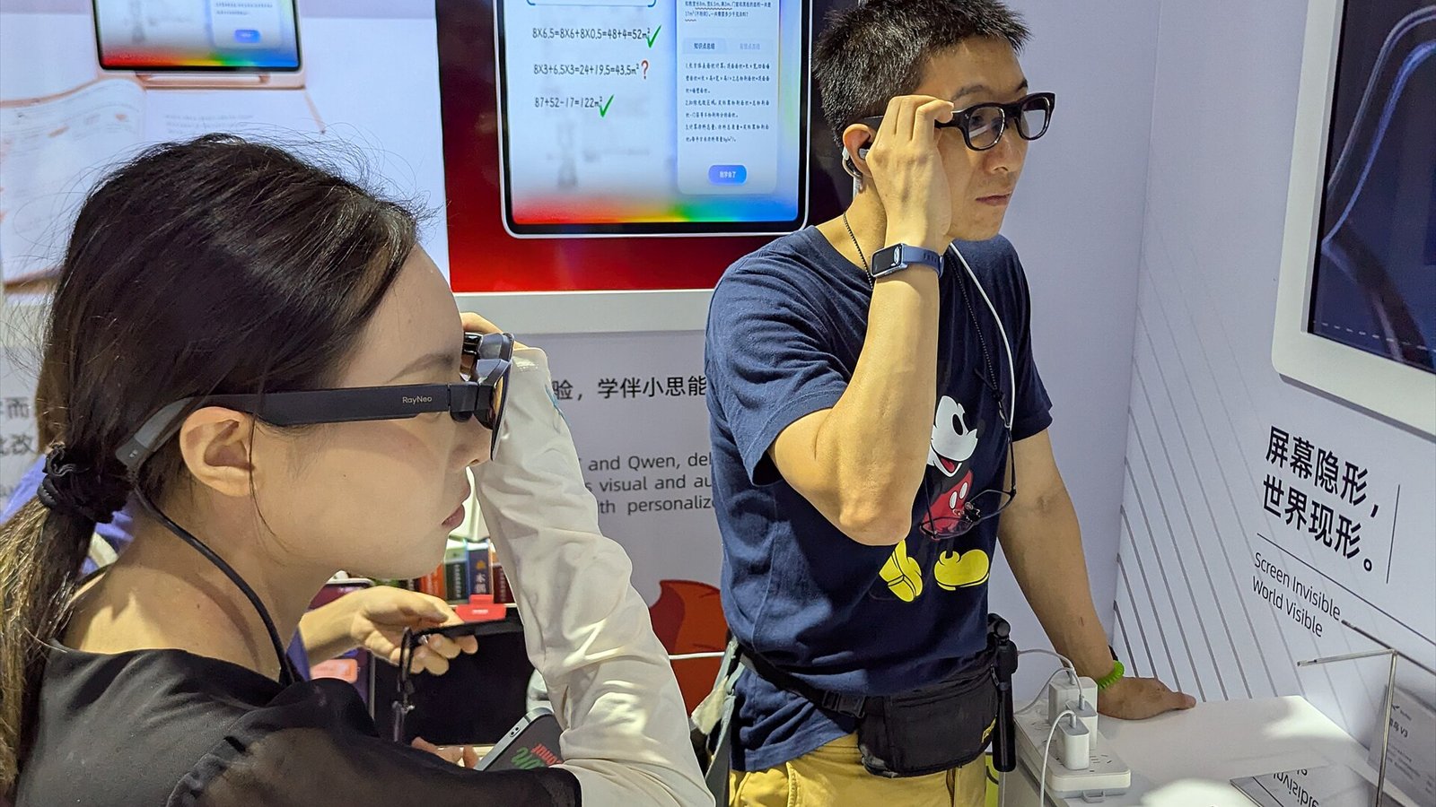 Two people demonstrate black smart glasses inside a technology exhibition booth.