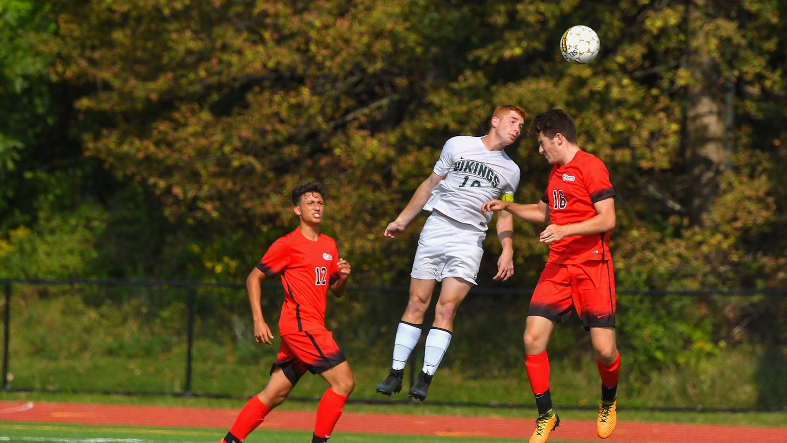 Three men's college soccer players watch the ball after one jumps for a header on a grass field.