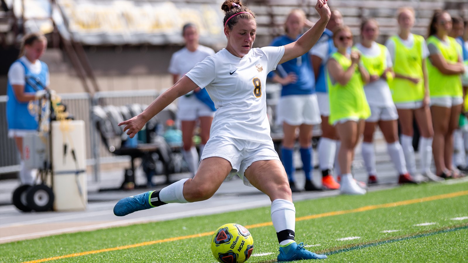 A women's soccer player in a white uniform swings her right foot through the ball near the touchline.