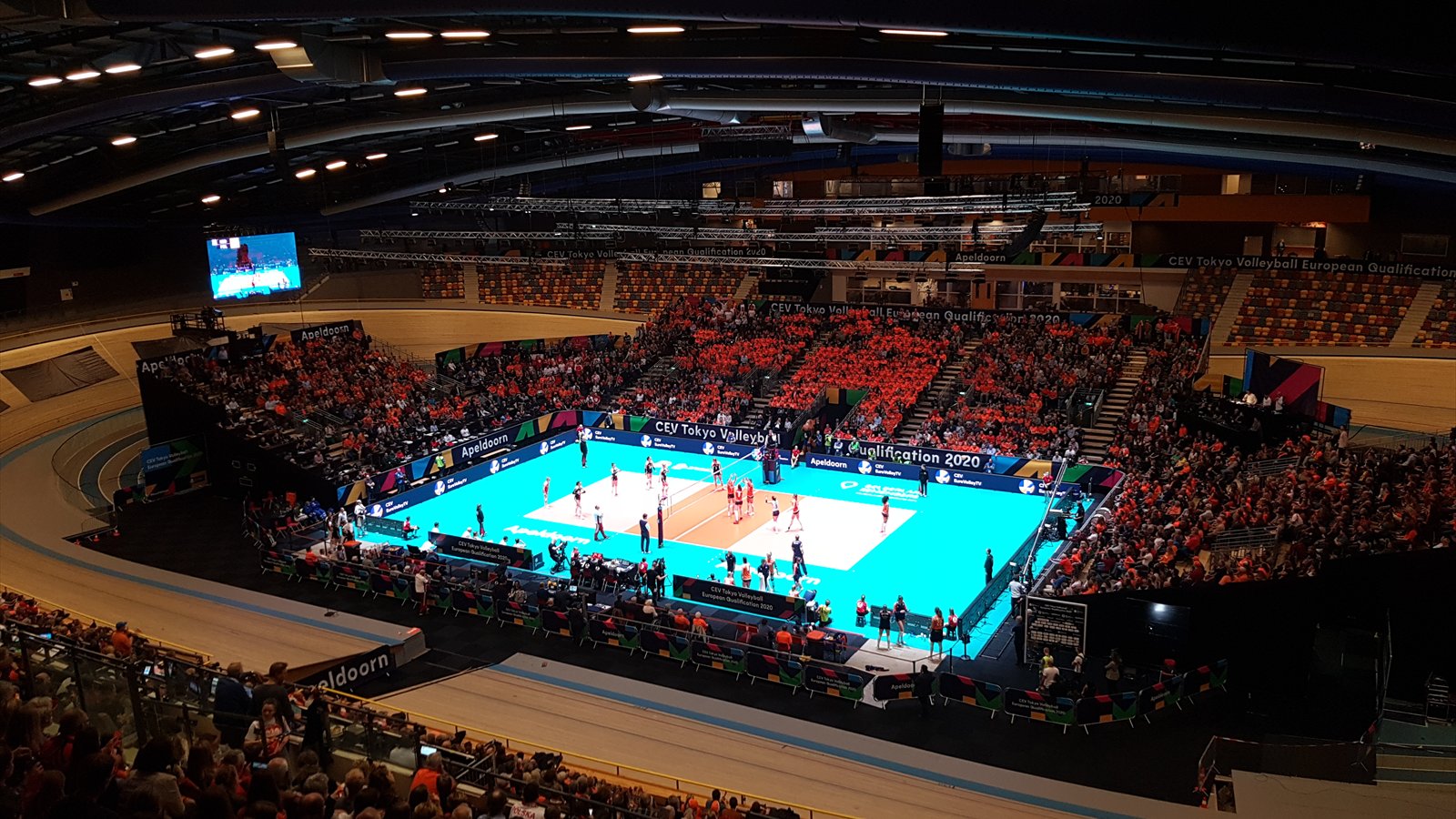 Women's volleyball teams compete on an indoor court before a large crowd.