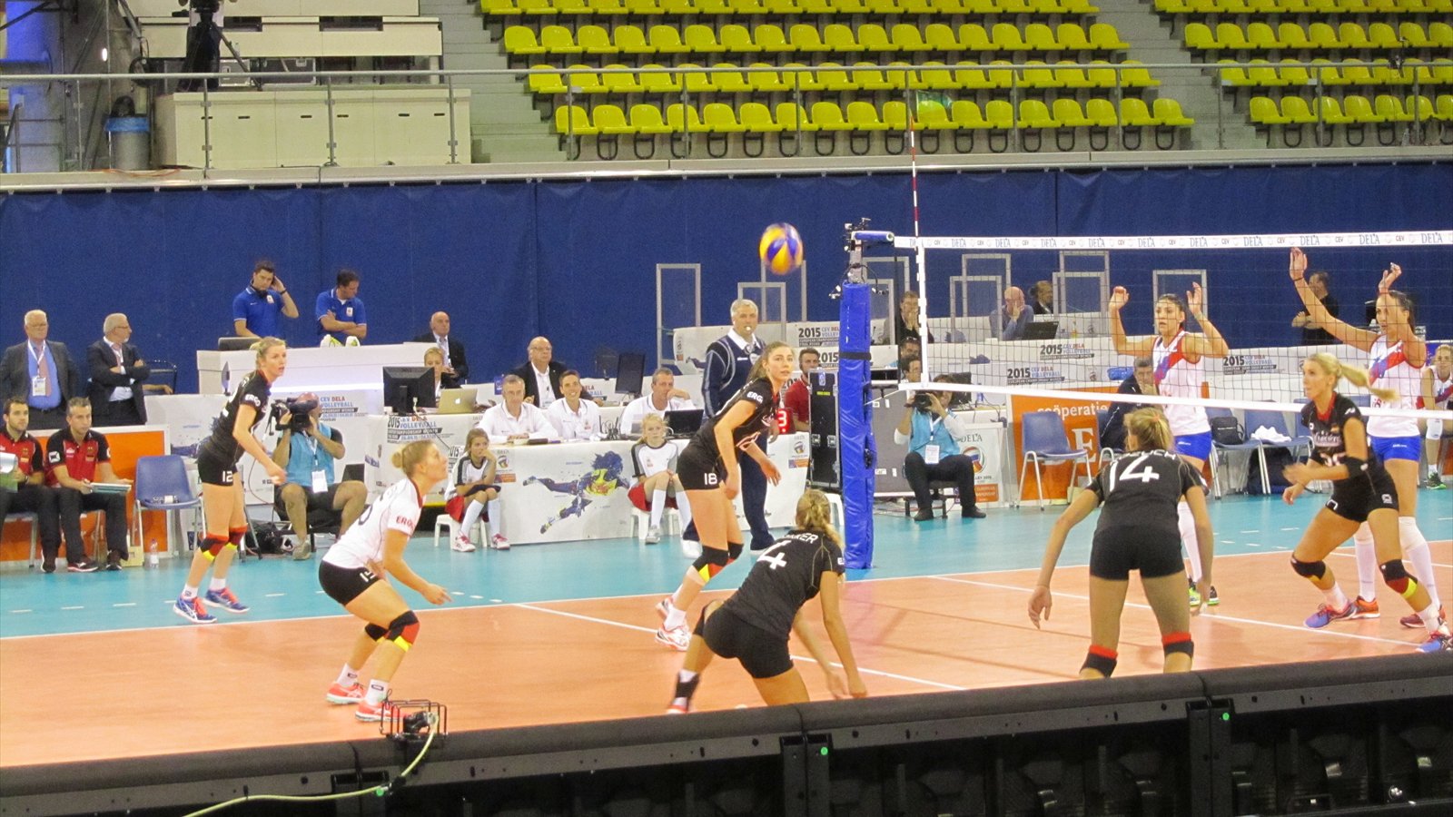 Women's volleyball players line up at the net during an indoor match as teammates watch from the back court.