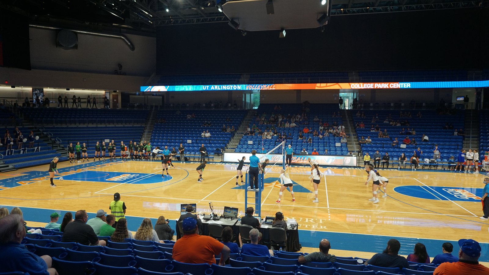 Women's college volleyball players track the ball across the net during an indoor match.