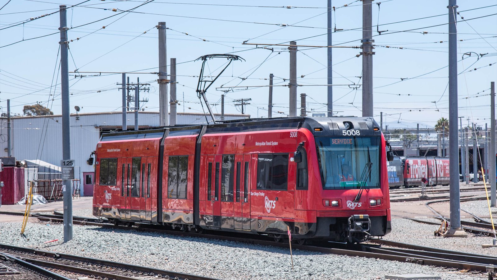 A red San Diego MTS trolley moves through a rail yard beneath overhead electrical wires.