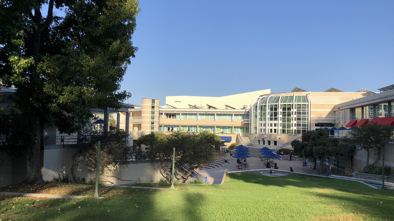 UC San Diego's Price Center buildings surround a sunlit lawn and lower plaza with trees and blue umbrellas.
