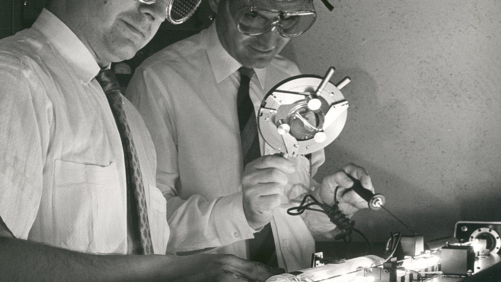 Two researchers wearing protective glasses work beside a glowing laser beam and optical equipment in a laboratory.