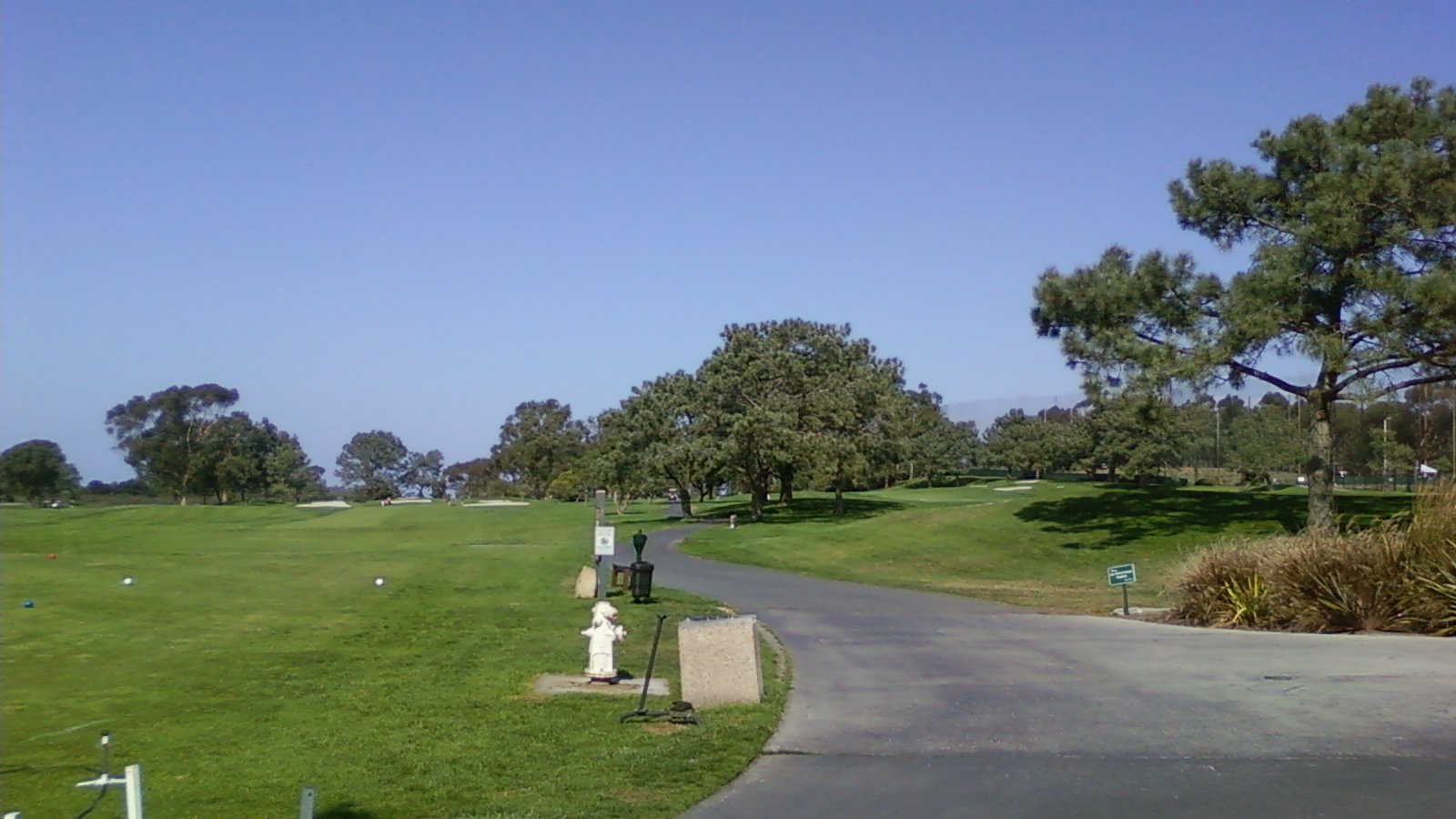 A paved cart path curves between green fairways and mature trees at Torrey Pines Golf Course beneath a clear blue sky.